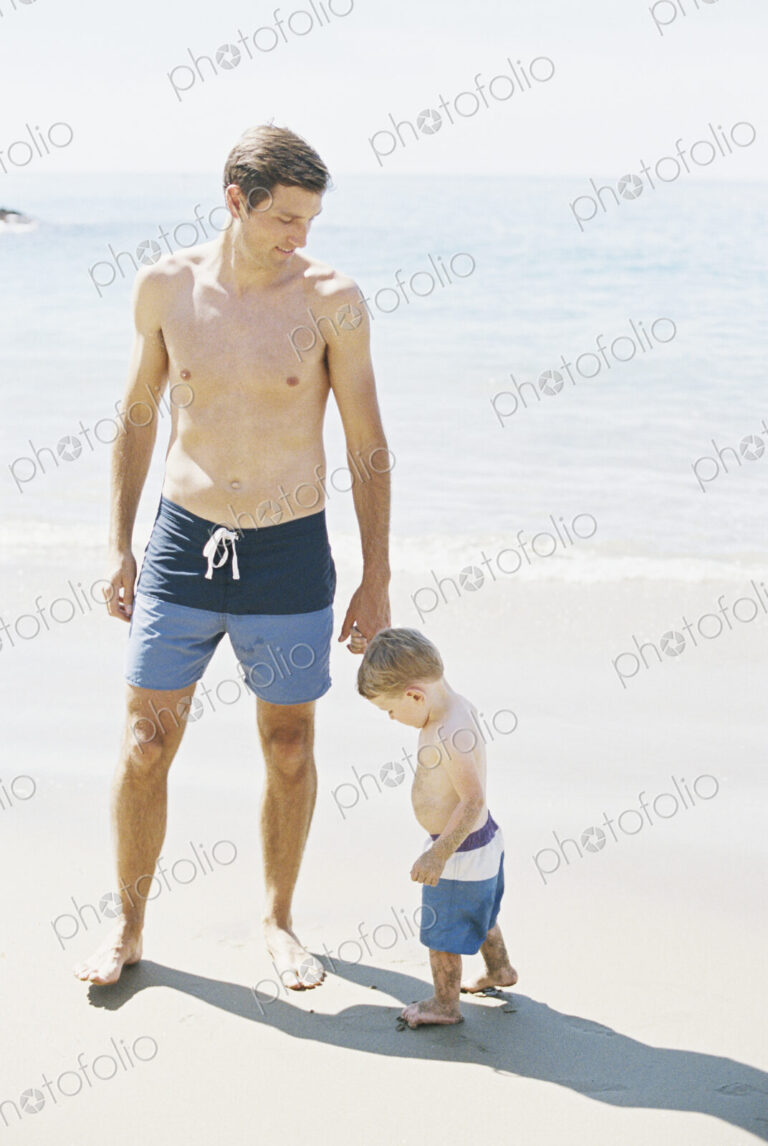 Man and his son wearing swimming trunks standing on a sandy beach by the ocean.