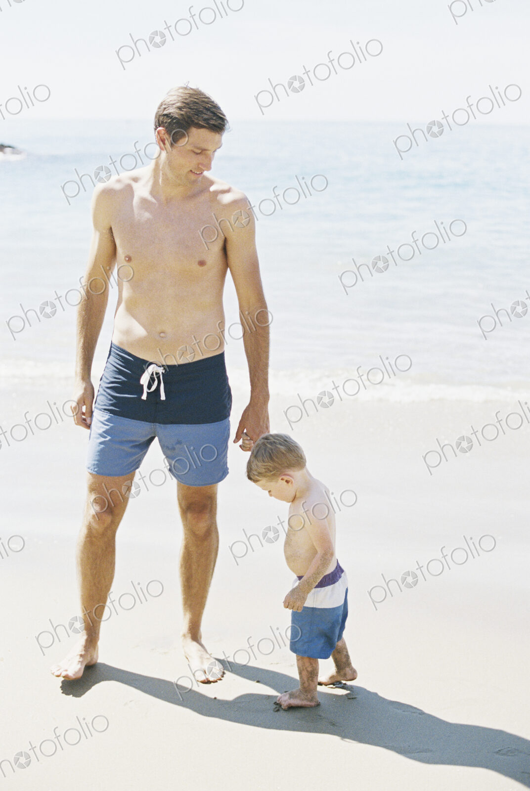 Man and his son wearing swimming trunks standing on a sandy beach by the ocean.