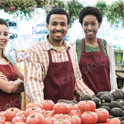 Man and two women wearing aprons standing at stall with fresh tomatoes at a fruit and…