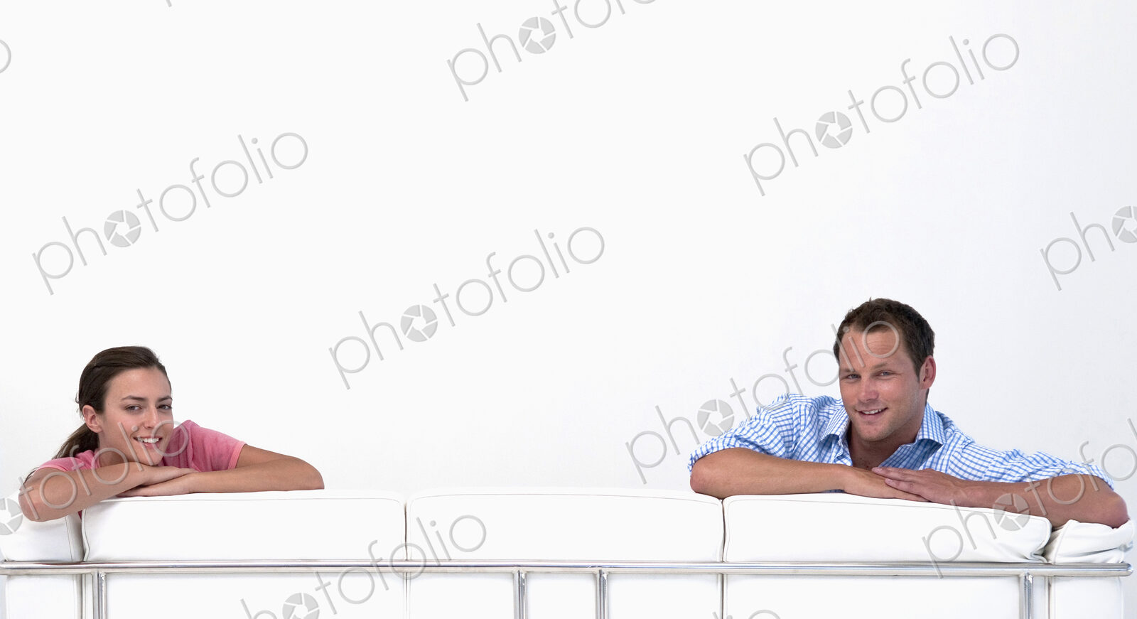 Man and woman looking over the back of a sofa at camera with white background.