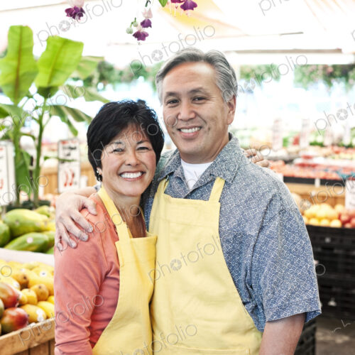 Man and woman owners of a fruit and vegetable market, wearing aprons, standing side by…
