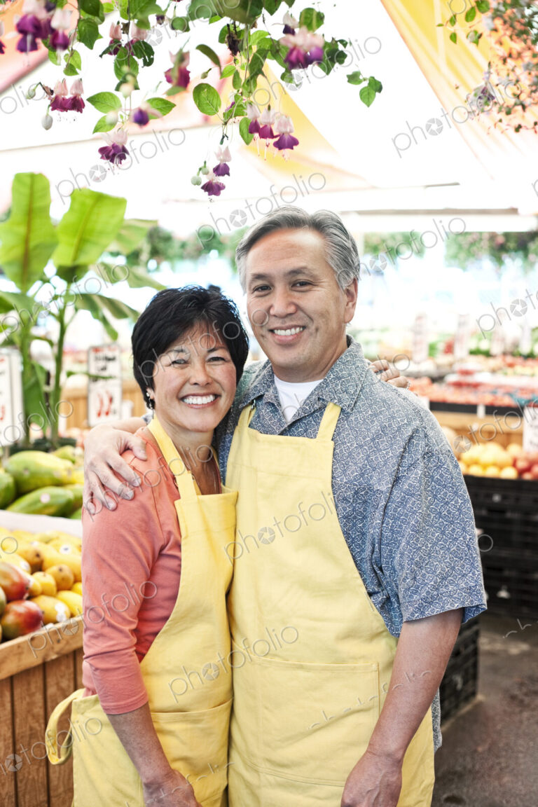 Man and woman owners of a fruit and vegetable market, wearing aprons, standing side by…