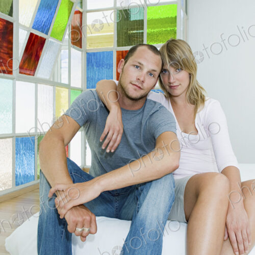 Man and woman sitting hugging on a futon bed by colourful glass windows. Man and woman sitting hugging on a futon bed by colourful glass windows.