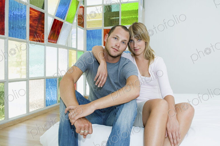 Man and woman sitting hugging on a futon bed by colourful glass windows.