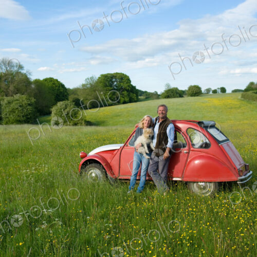 Man and woman standing side by side on a meadow, holding small dog, leaning against…