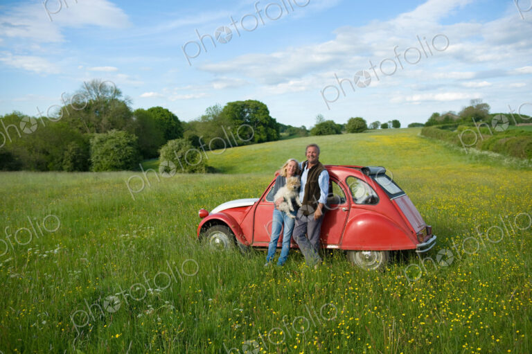 Man and woman standing side by side on a meadow, holding small dog, leaning against…