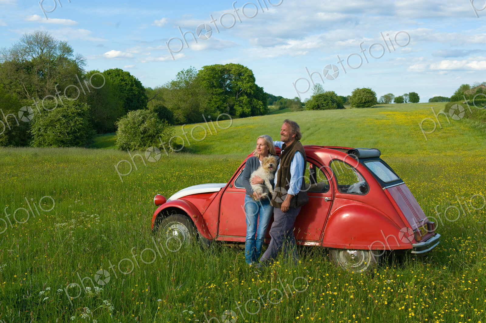 Man and woman standing side by side on a meadow, holding small dog, leaning against…