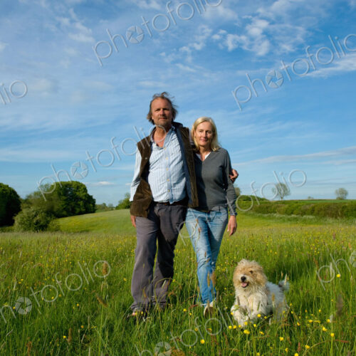 Man and woman walking arm in arm across a meadow, small dog running beside them.