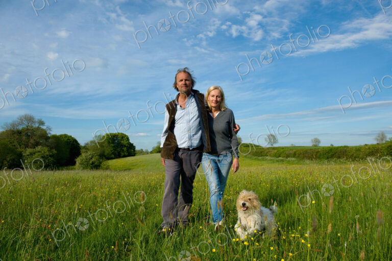 Man and woman walking arm in arm across a meadow, small dog running beside them.