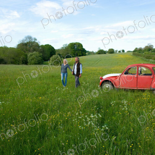 Man and woman walking hand in hand across a meadow, vintage red car parked close by.