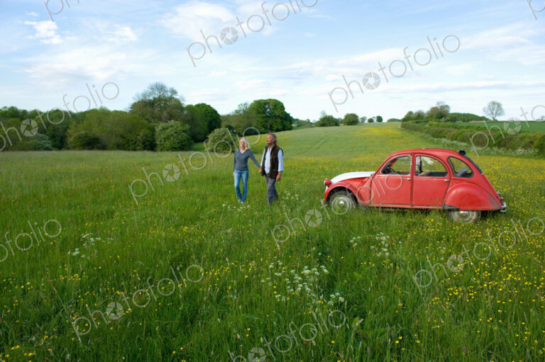 Man and woman walking hand in hand across a meadow, vintage red car parked close by.