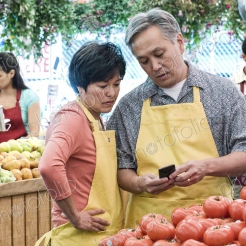 Man and woman wearing aprons standing at stall with fresh tomatoes at a fruit and…