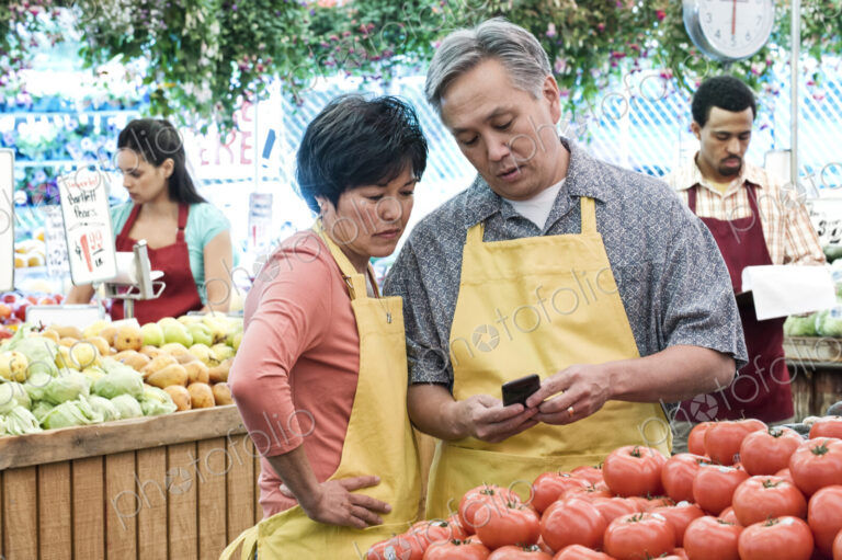 Man and woman wearing aprons standing at stall with fresh tomatoes at a fruit and…