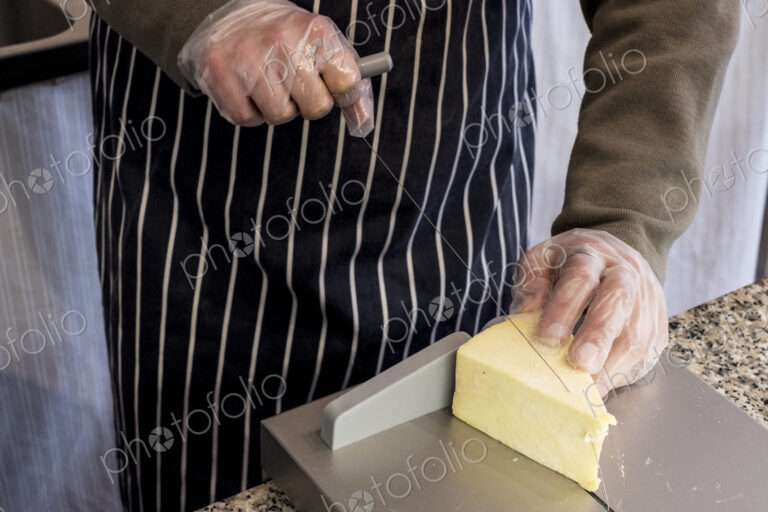 Man at counter in a delicatessen, slicing cheese with wire cutter.