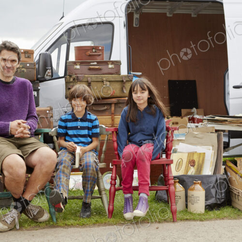 Man, boy and girl sitting on vintage chairs in front of a bric-a-brac stall with…