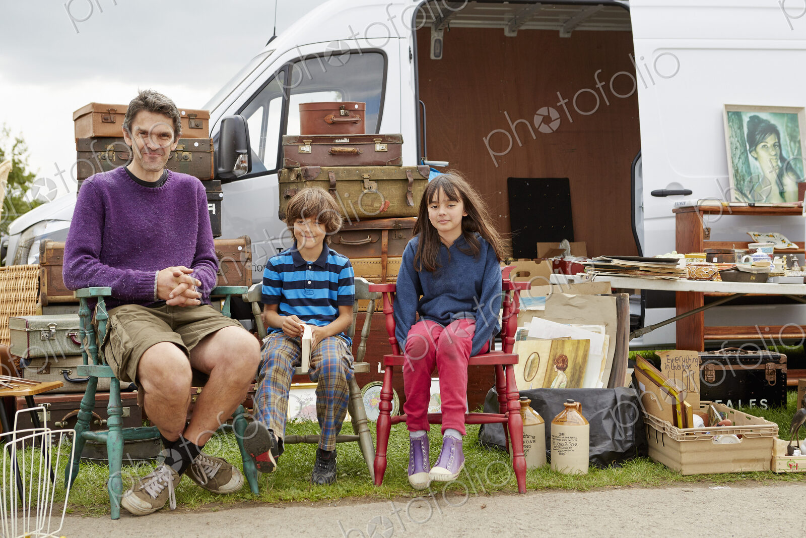 Man, boy and girl sitting on vintage chairs in front of a bric-a-brac stall with…