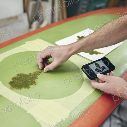 Man creating a design on surfboard laying an oak leaf on the board, using mobile phone…