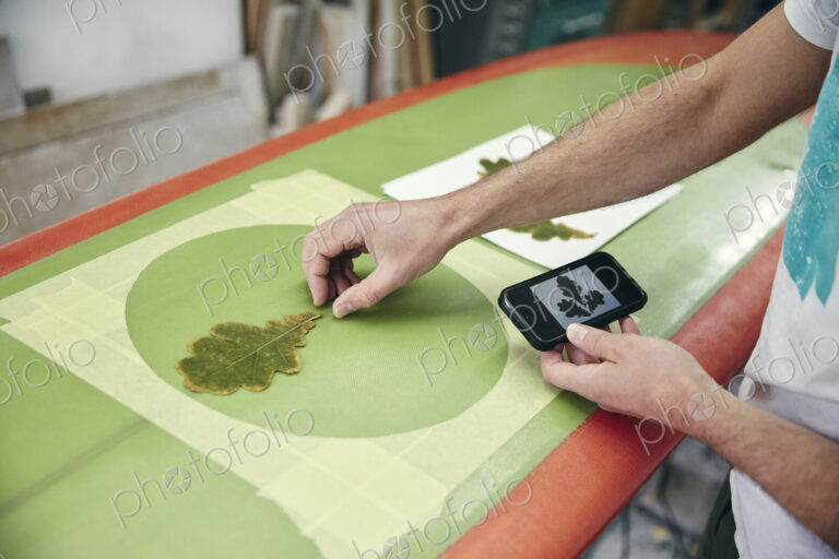 Man creating a design on surfboard laying an oak leaf on the board, using mobile phone…