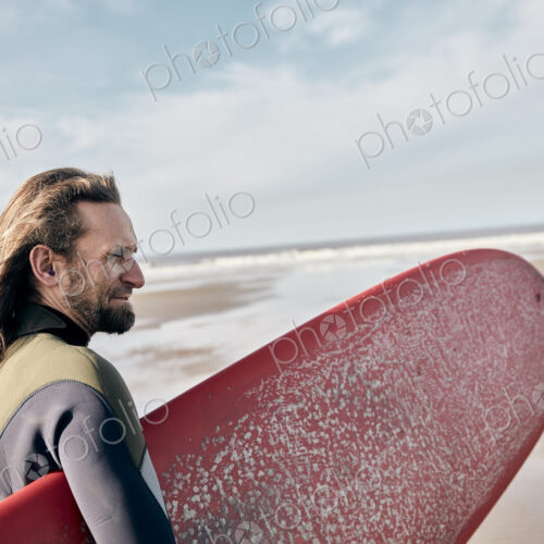 Man holding a full size surf board standing on beach looking out to sea Man holding a full size surf board standing on beach looking out to sea