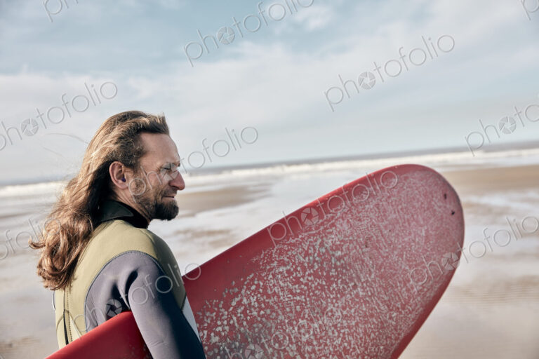 Man holding a full size surf board standing on beach looking out to sea