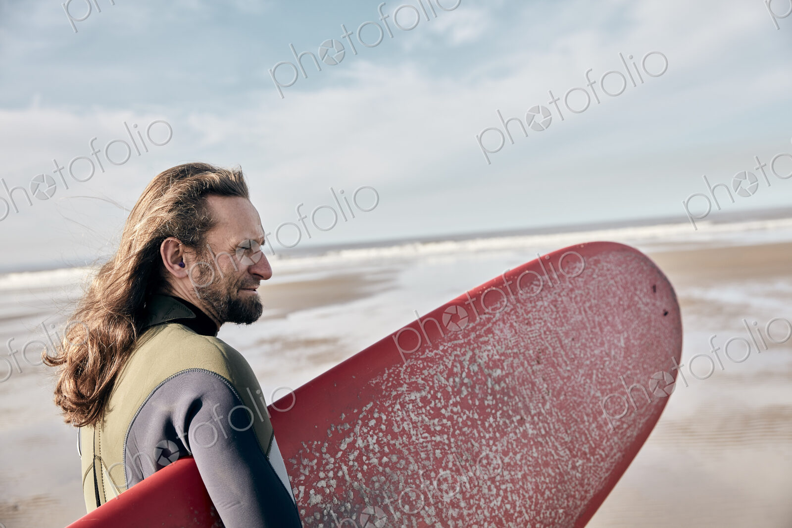 Man holding a full size surf board standing on beach looking out to sea