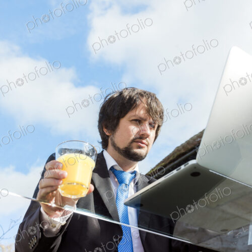 Man holding glass of juice working on a glass desk outside.