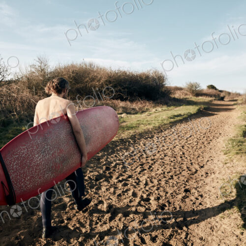 Man holding surf board walking along sandy path towards beach Man holding surf board walking along sandy path towards beach