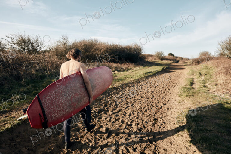 Man holding surf board walking along sandy path towards beach