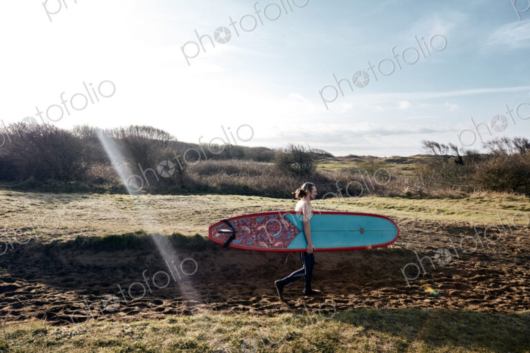 Man holding surf board walking along sandy path towards the sea