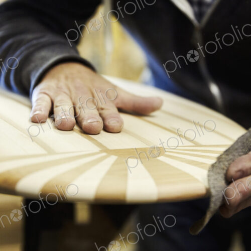 Man in a workshop sanding the edge of a wooden surfboard with a surface pattern of… Man in a workshop sanding the edge of a wooden surfboard with a surface pattern of…