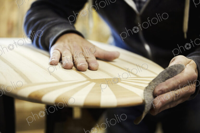 Man in a workshop sanding the edge of a wooden surfboard with a surface pattern of…