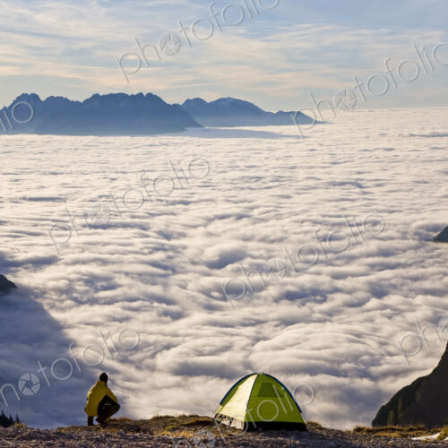 Man kneeling next to tent pitched above the clouds on a mountain summit.
