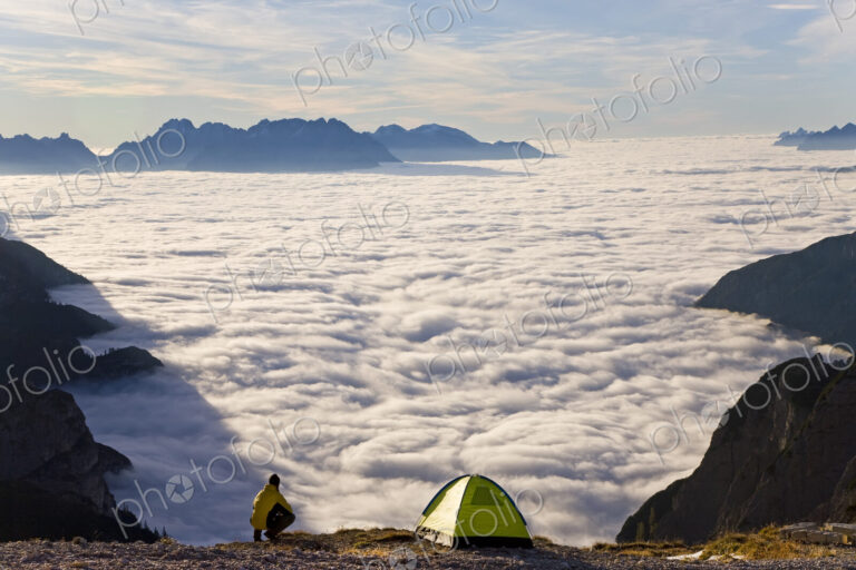 Man kneeling next to tent pitched above the clouds on a mountain summit.