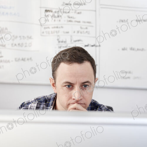 Man looking at a computer screen seated at his desk, with a project chart on the…
