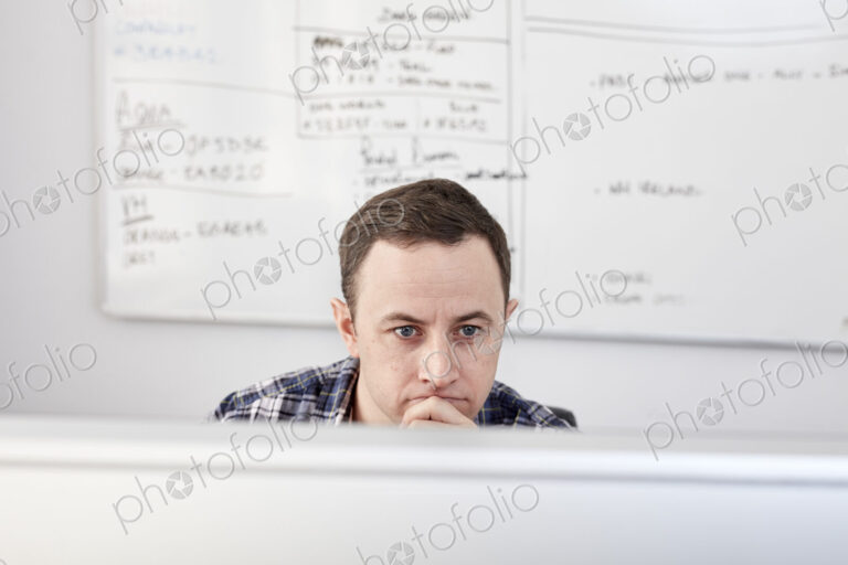Man looking at a computer screen seated at his desk, with a project chart on the…