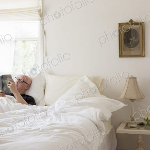 Man lying in a bed with white linen, reading.