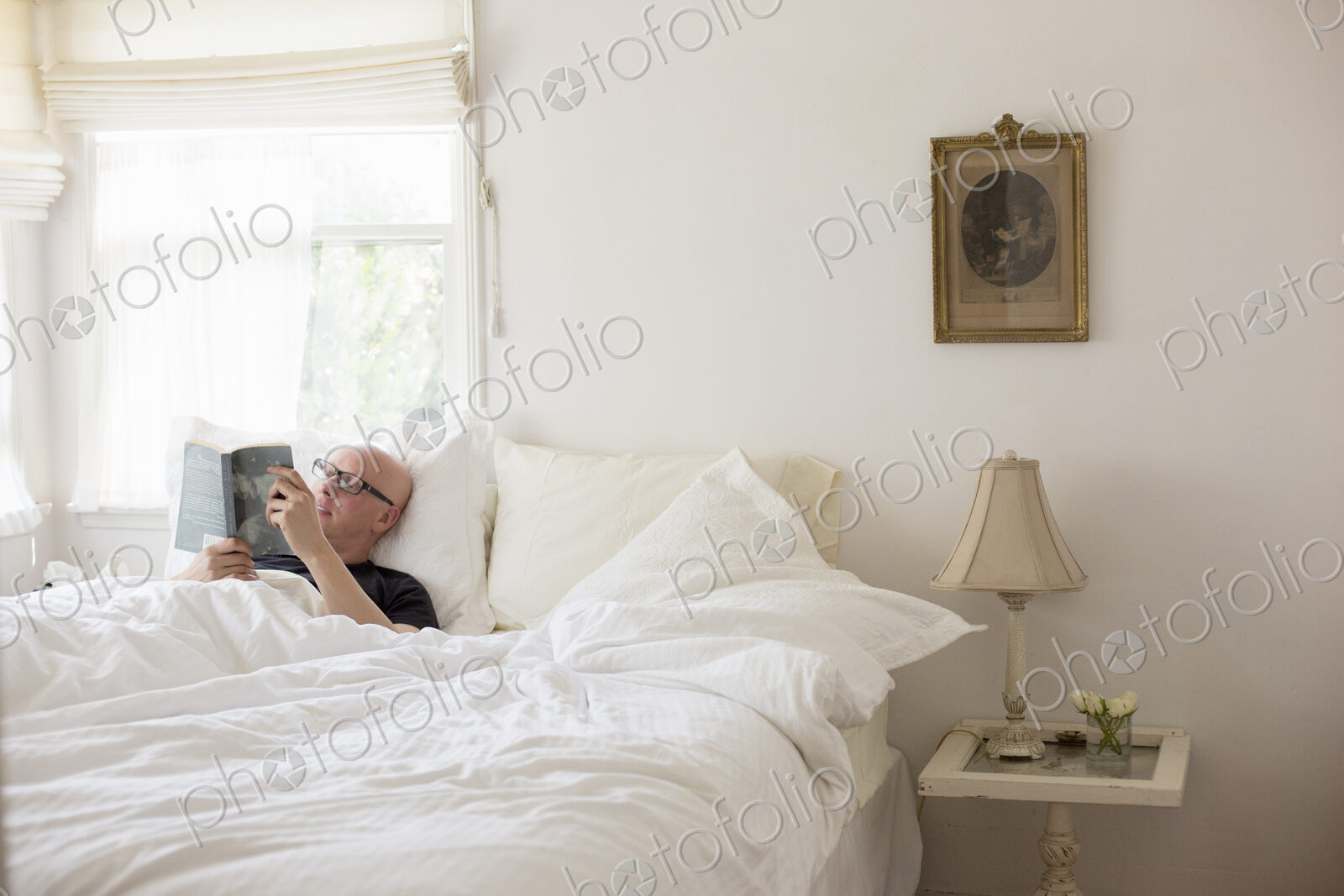 Man lying in a bed with white linen, reading.