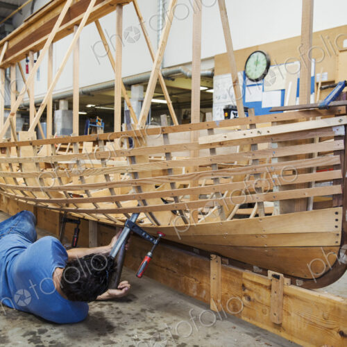 Man lying on floor in a boat-builder's workshop, working on a wooden boat hull.