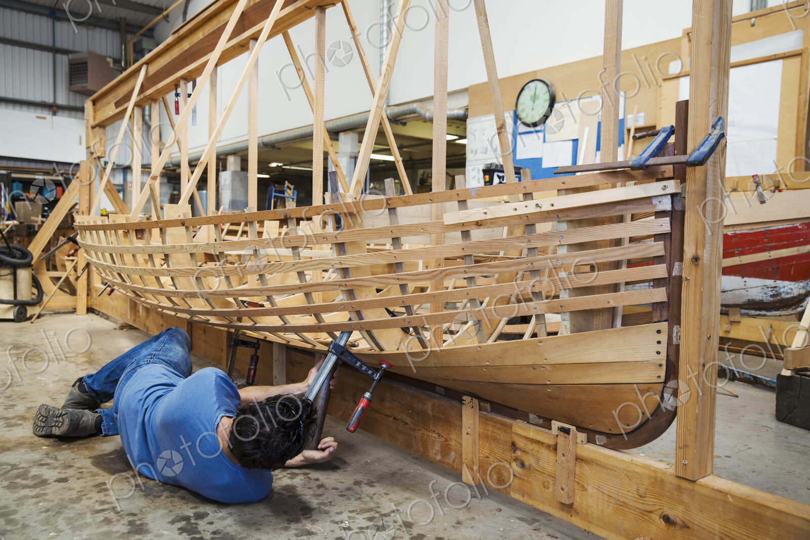 Man lying on floor in a boat-builder’s workshop, working on a wooden boat hull.