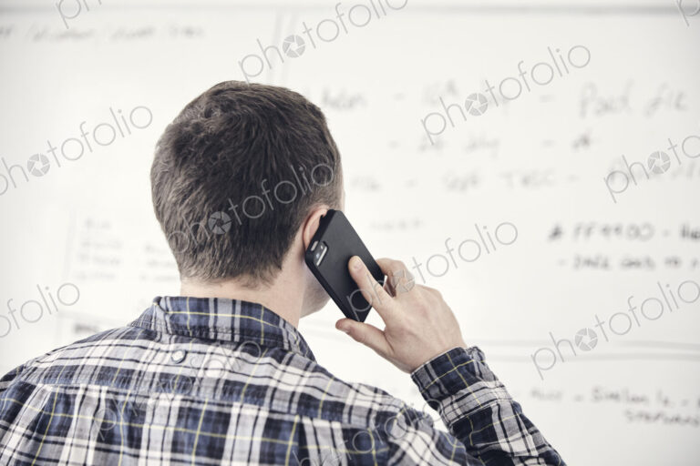 Man on mobile phone with his back to the camera looking at a whiteboard in an office.