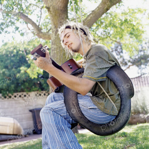 Man playing guitar sitting on a tyre swing in a garden, singing.