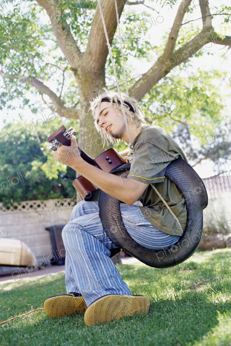 Man playing guitar sitting on a tyre swing in a garden, singing.