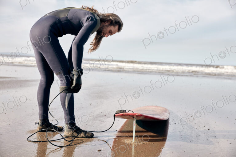 Man preparing to surf on a sandy beach in a wetsuit.