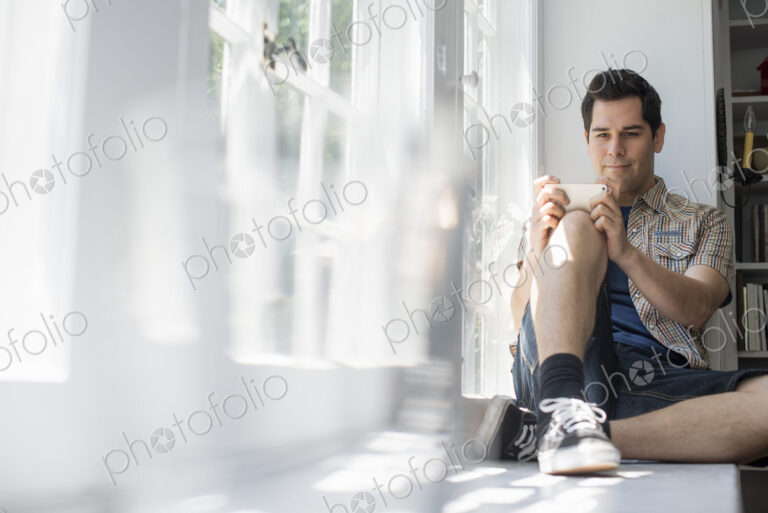 Man sitting by a window, holding a cell phone.