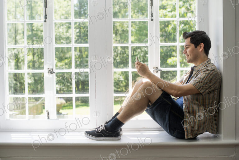 Man sitting by a window, holding a cell phone.