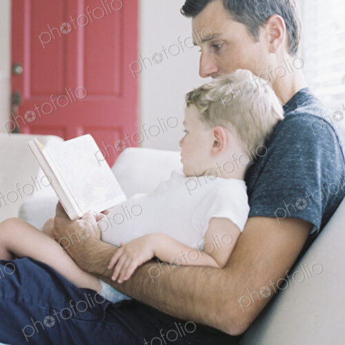 Man sitting on a sofa with his son sitting on his lap, holding a book, reading a story. Man sitting on a sofa with his son sitting on his lap, holding a book, reading a story.