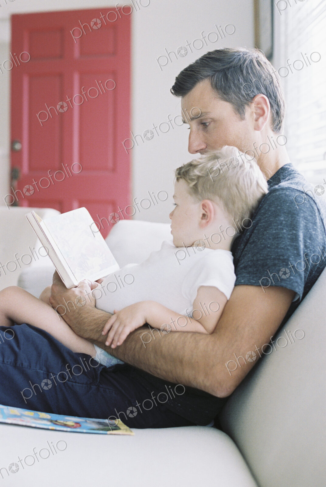 Man sitting on a sofa with his son sitting on his lap, holding a book, reading a story.