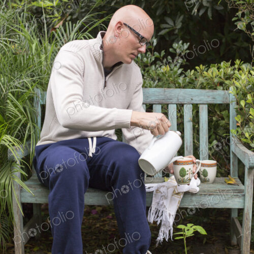 Man sitting on a wooden bench in a garden, pouring a cup of coffee. Man sitting on a wooden bench in a garden, pouring a cup of coffee.