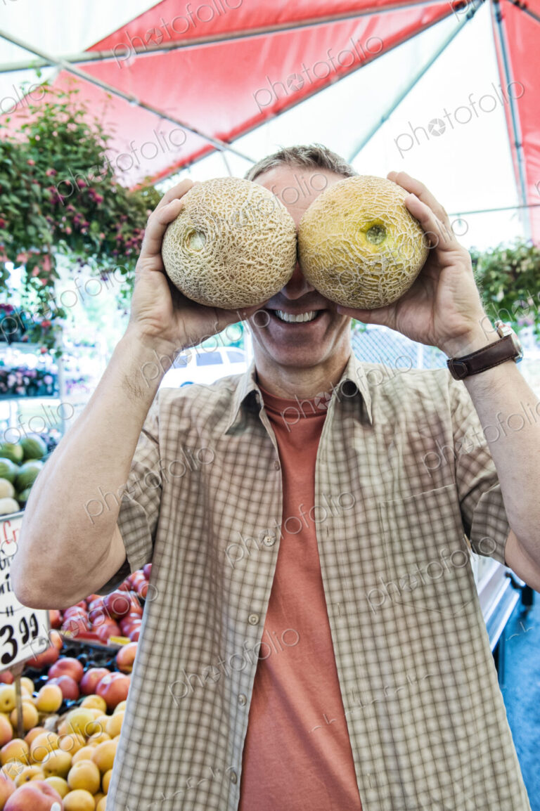 Man standing at a farm stall, covering his eyes with two fresh melons, smiling at camera.