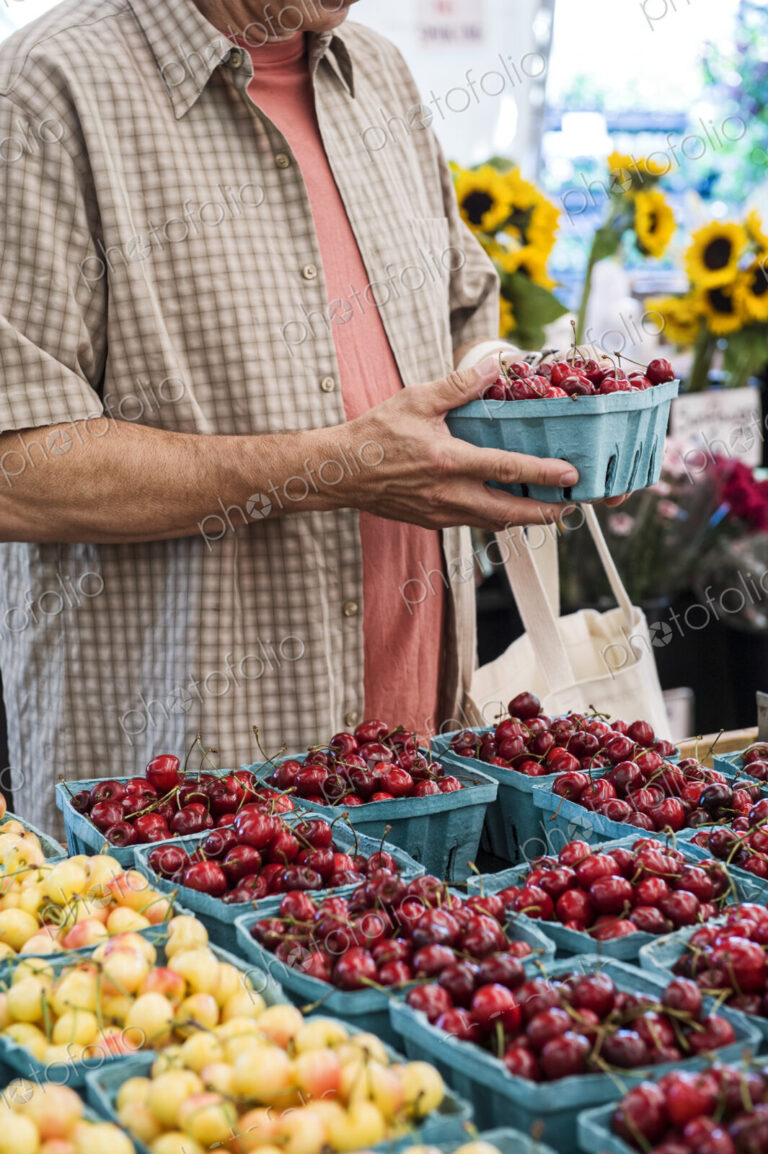 Man standing at stall, holding punnet with fresh cherries at a fruit and vegetable market.